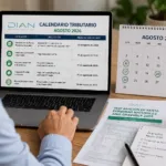 Persona revisando el calendario tributario de agosto de 2026 en un computador portátil, con documentos de impuestos y el calendario de vencimientos de la DIAN sobre un escritorio.